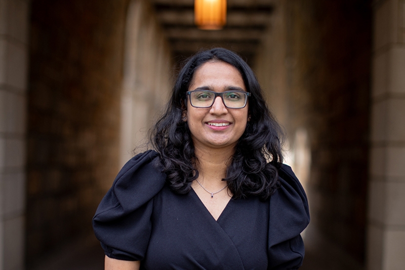 A person with shoulder length hair smiles in front of a stone archway.
