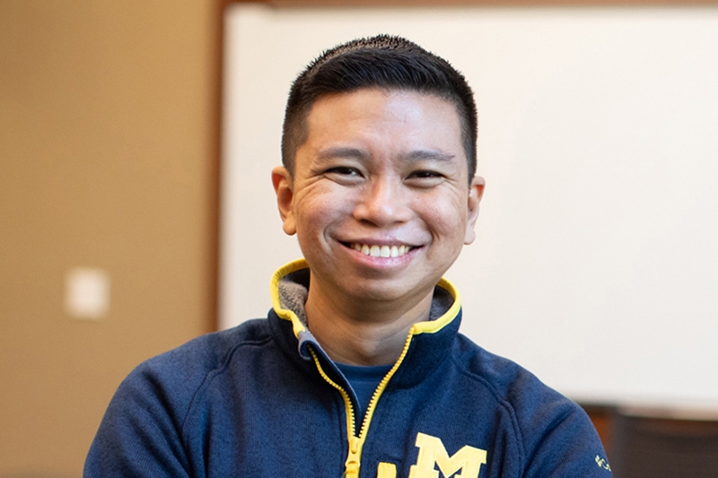A person with short hair smiles in front of a classroom markerboard.
