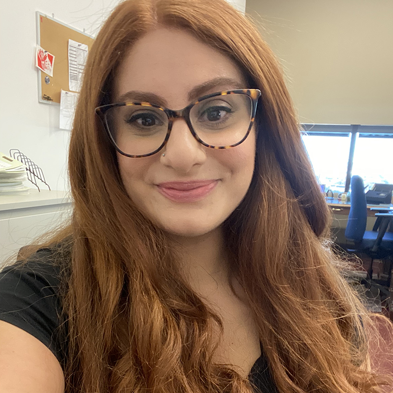 A woman with long auburn hair and glasses smiles in an office. 