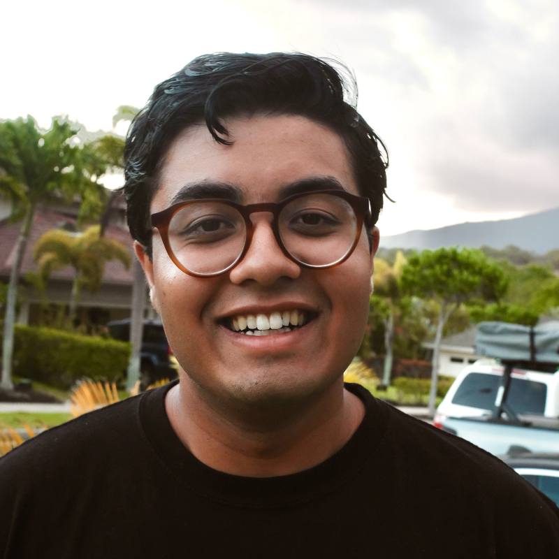 A person with short hair and glasses smiles in front of palm trees and a cloudy sky.