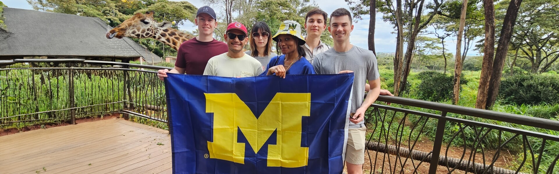 Michigan law students holding up a University of Michigan flag in South Africa, with a giraffe in the background grazing from a tree.