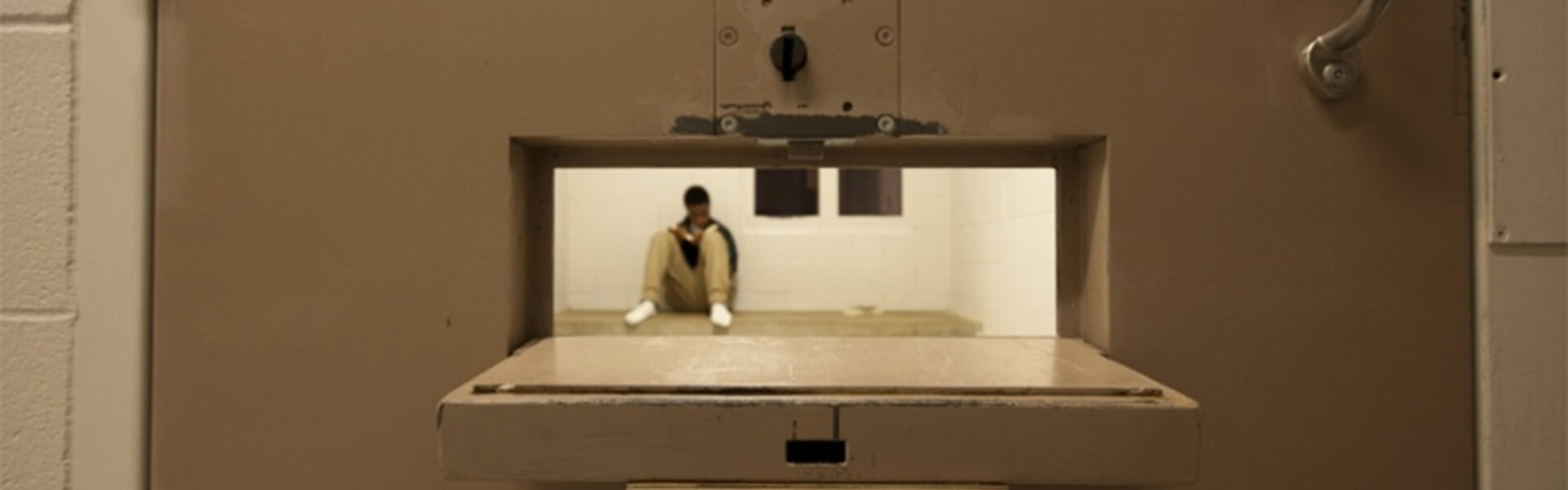 Looking though the food pass door of a jail cell with an inmate sitting on the floor.