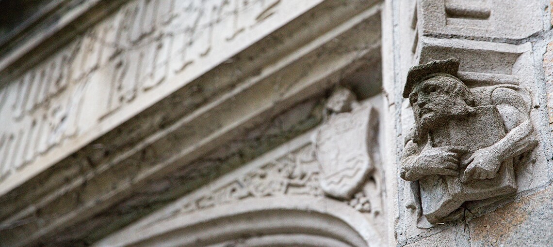 Architectural close up of stone work on the outside of the Quadrangle. A scholar holding a book. 