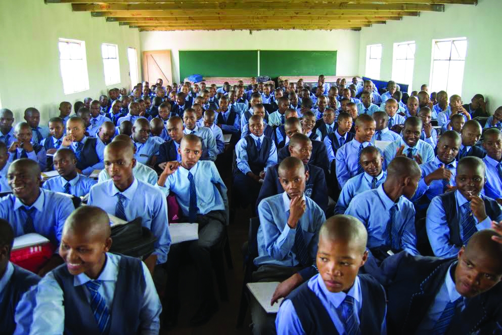 The interior of a classroom in South Africa, where dozens of male students wearing shirts and ties are packed shoulder-to-shoulder.