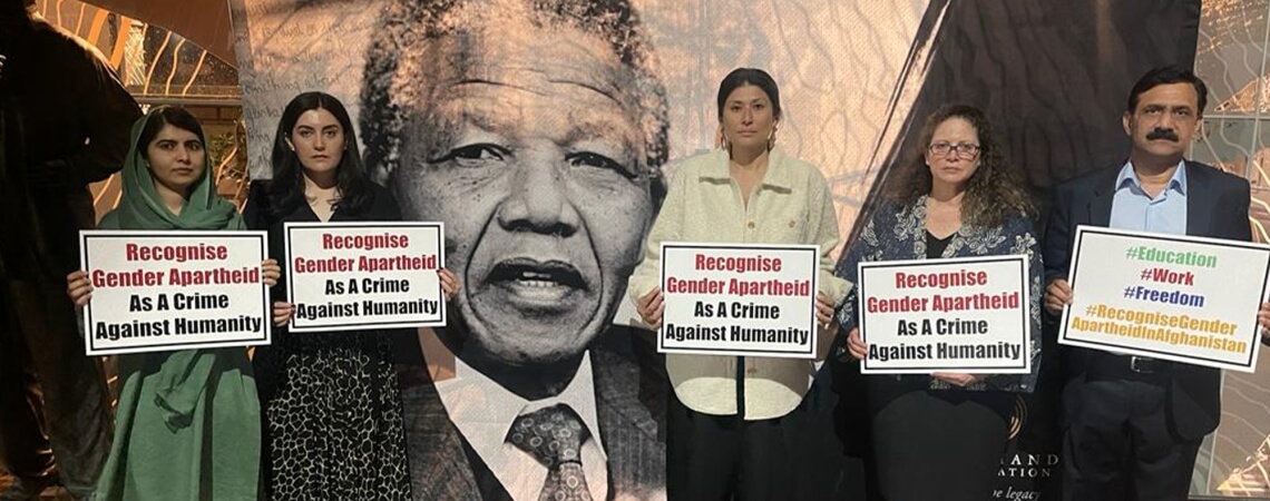 Five people stand in solidarity, holding signs that demand recognition of gender apartheid as a crime against humanity at the Nelson Mandela Annual Lecture