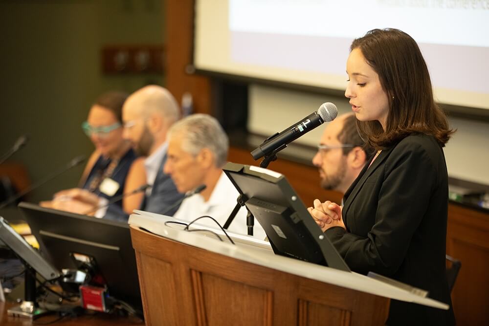 A woman in a suit stands at a lectern addressing the conference attendees.