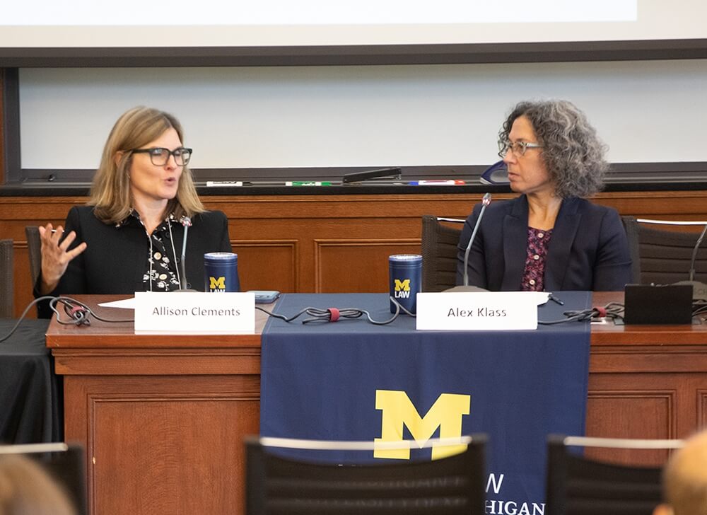 Two women sit behind a wood panel desk mid-discussion.