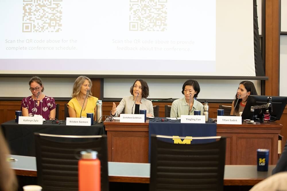Panelists sit behind a wooden desk speaking to a conference audience.