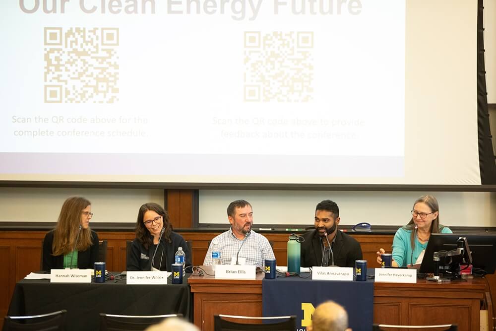 Panelists sit behind a wooden desk speaking to a conference audience.