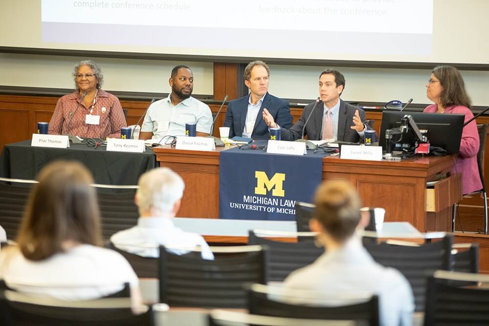 Panelists sit behind a wooden desk speaking to a conference audience.
