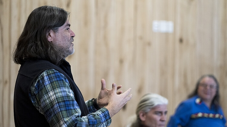 A person with shoulder-length hair, a beard, and a flannel shirt speaks to a group of students.