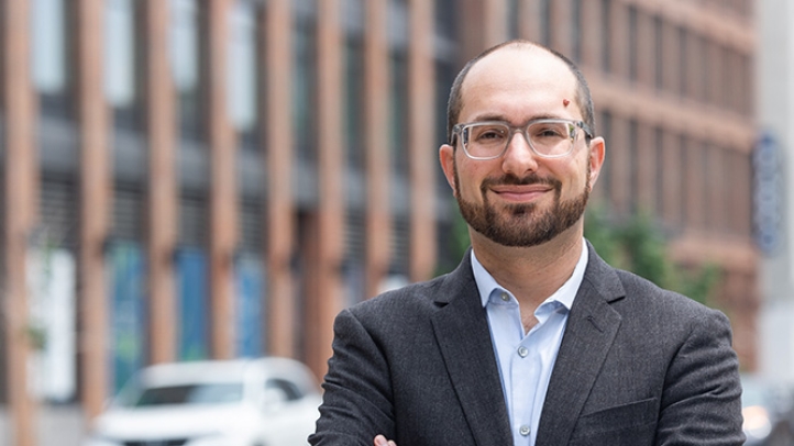 Professor Noah Kazis looking at camera with a blurry city building behind him.