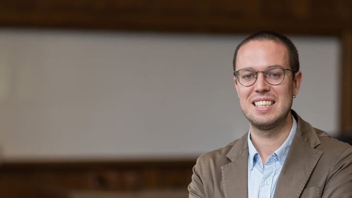 Man, standing at the front of a classroom with a large whiteboard behind him. He is smiling and looking friendly.