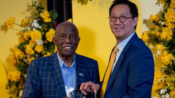 Two people standing in front of a festive background. One person is handing a leadership award to the other person.  