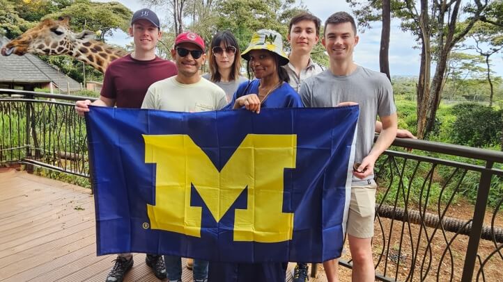 Five individuals stand on a balcony, proudly holding a University of Michigan flag, with a giraffe in the background.