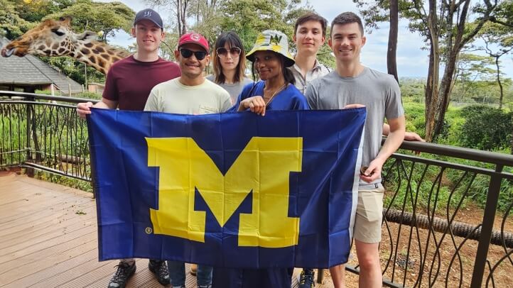 Five individuals stand on a balcony, proudly holding a University of Michigan flag, with a giraffe in the background.