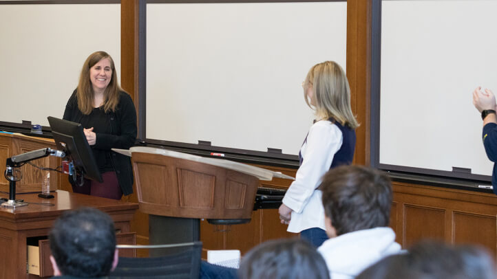 A professor stands behind a lectern while two others stand and clap for her.