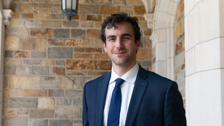 A man in a suit smiles in front of a stone wall.