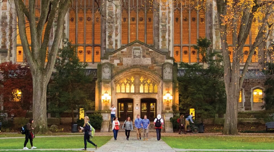Students walking through the quad at dusk with the reading room glowing behind them.
