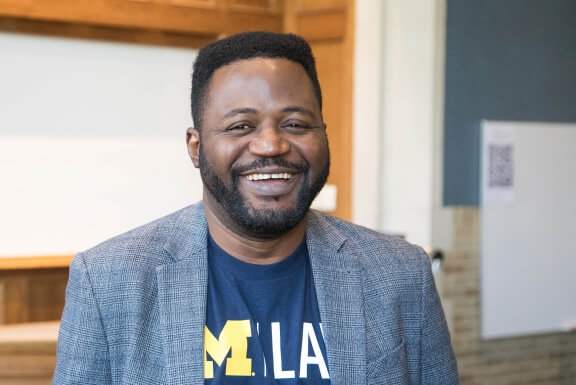 Rotimi Adejoorin smiles and leans against a Michigan Law classroom table.