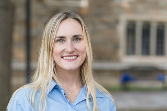 A woman with long blonde hair and a button down shirt smiles in front of a stone building.