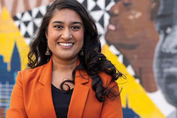 A woman with long dark hair and a bright blazer smiles in front of a colorful mural. 