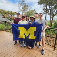 Five individuals stand on a balcony, proudly holding a University of Michigan flag, with a giraffe in the background.